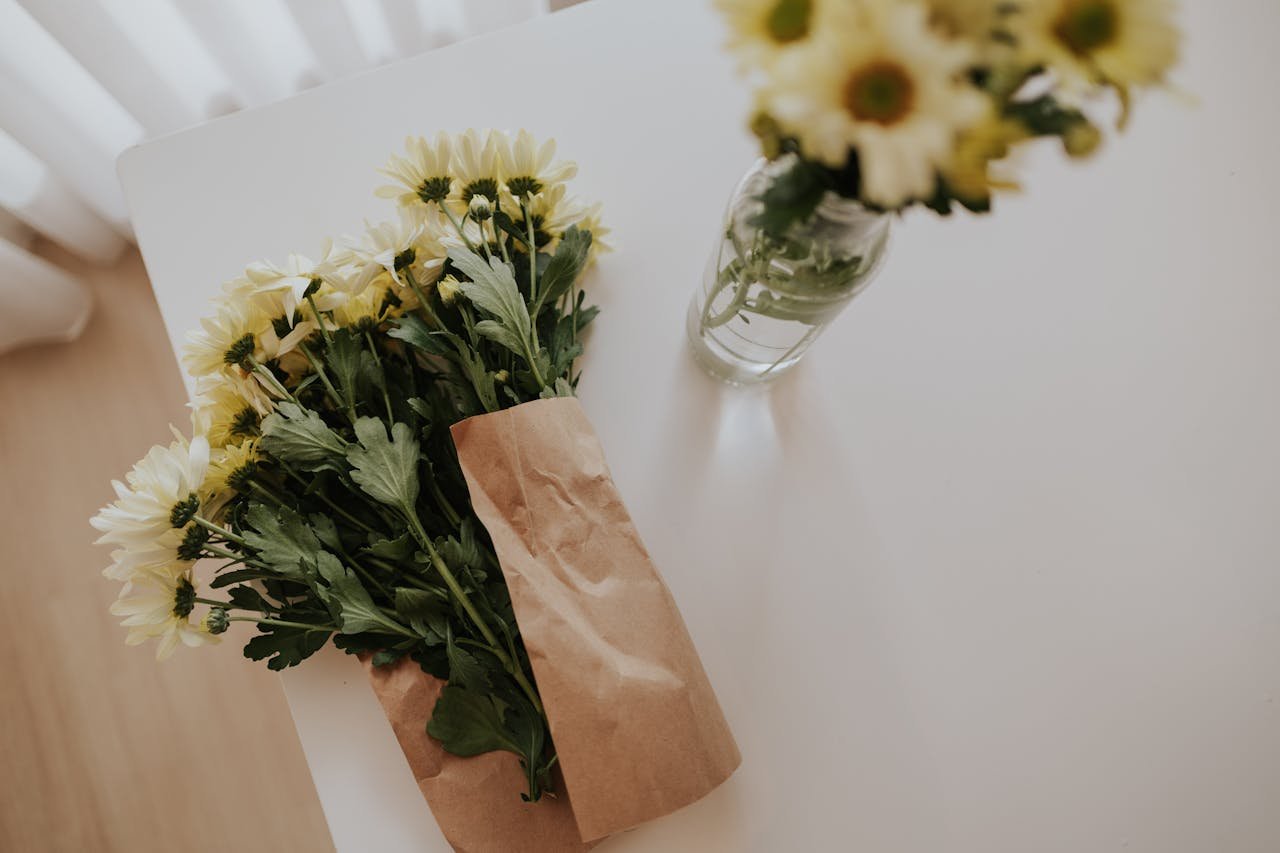 Flat lay of yellow chrysanthemums in paper wrap and vase on a clean white table.