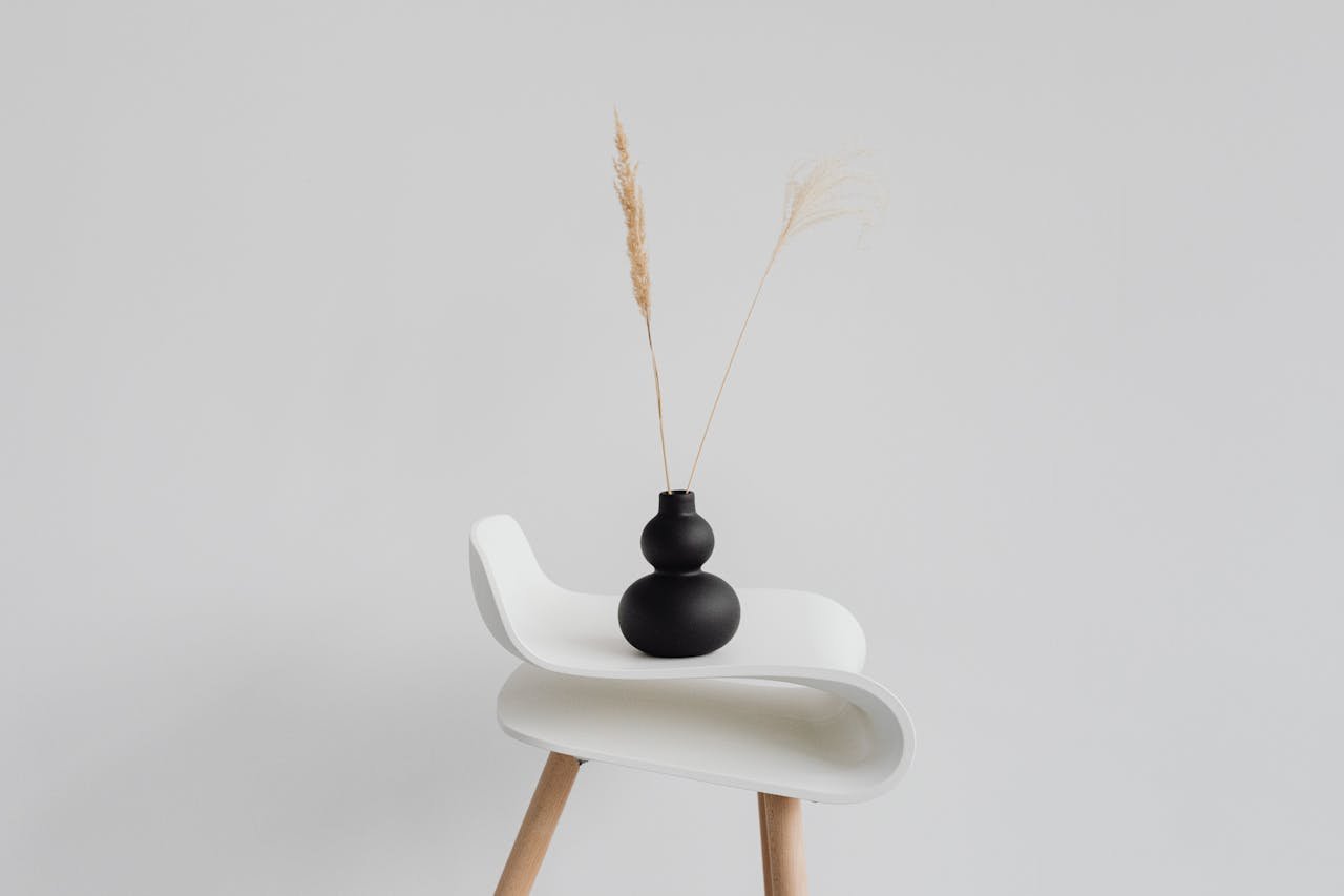 A sleek black vase with wheat ears on a modern white chair against a minimal background.